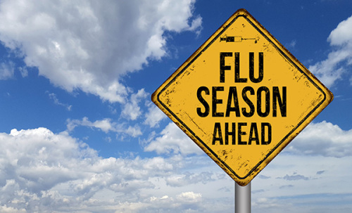 A yellow, diamond shaped road sign with the words Flu Season Ahead painted on it